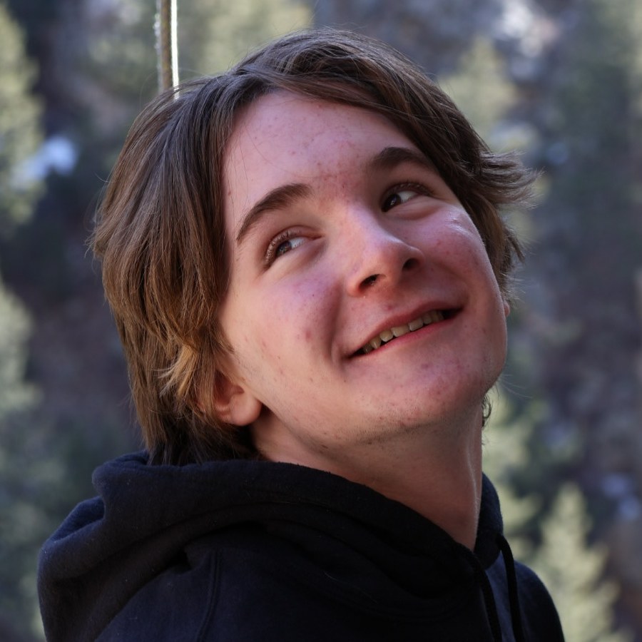 Thad outdoors in a black hoodie, looking up and smiling, pines and canyon rock behind him